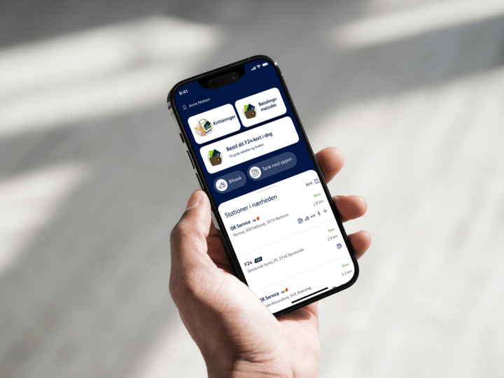 En hånd holder en smartphone med en app åben, der viser stationer i nærheden, kvitteringer og muligheder for at bestille f24-kort. Skærmen har en mørk blå baggrund med hvide og grønne elementer.